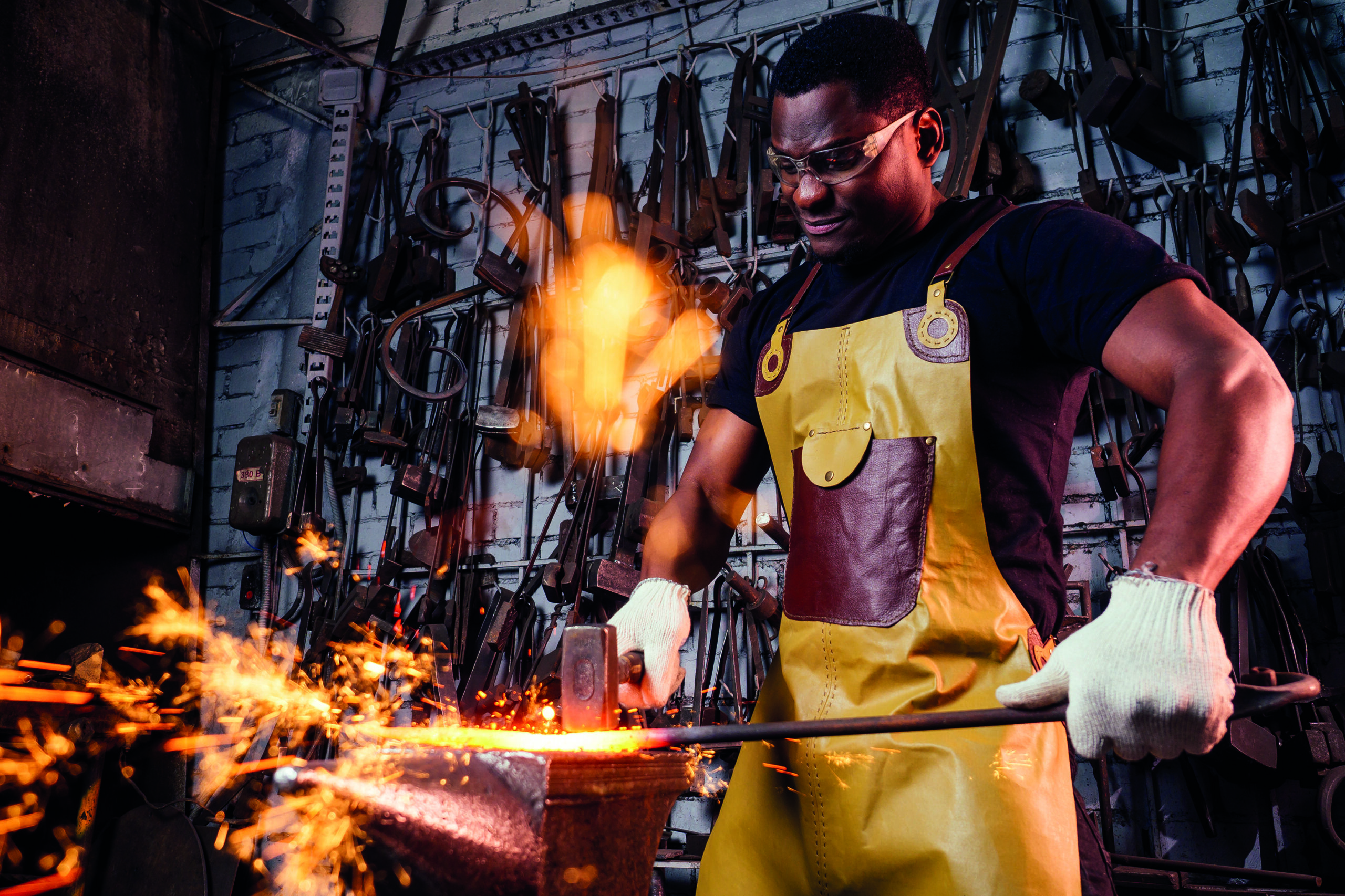 handsome african americam man forging steel next to furnace in dark workshop. small business comcept.